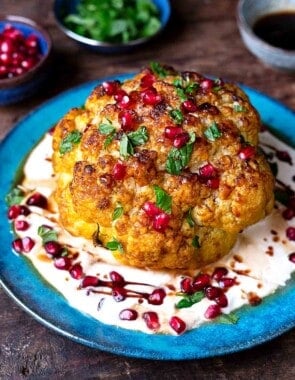 a whole roasted cauliflower with feta and yogurt sauce topped with pomegranate dressing and mint leaves on a plate.