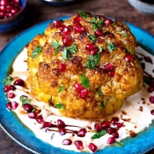 a whole roasted cauliflower with feta and yogurt sauce topped with pomegranate dressing and mint leaves on a plate.