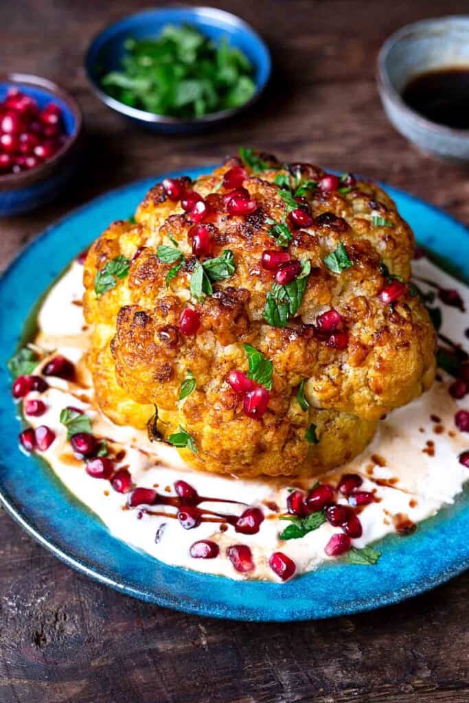 a whole roasted cauliflower with feta and yogurt sauce topped with pomegranate dressing and mint leaves on a plate.