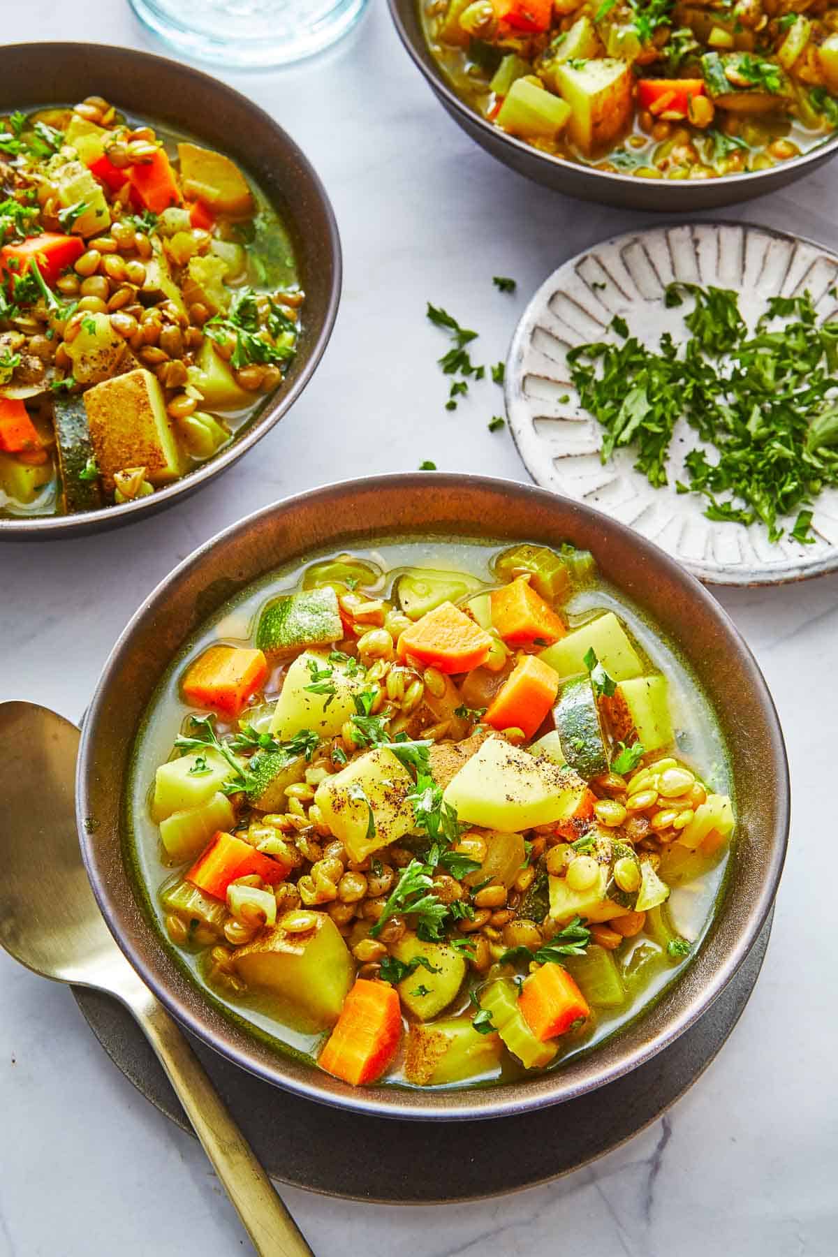 vegan lentil vegetable stew in three bowls next to a spoon and a bowl of chopped parsley.