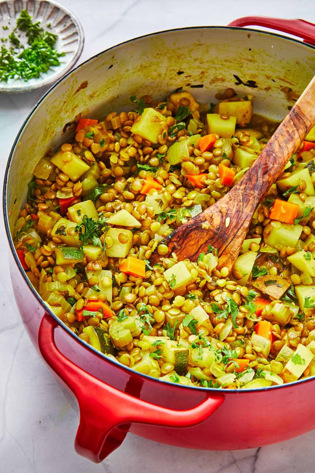 vegan lentil vegetable stew in a dutch oven with a wooden spoon, next to a bowl of chopped parsley.