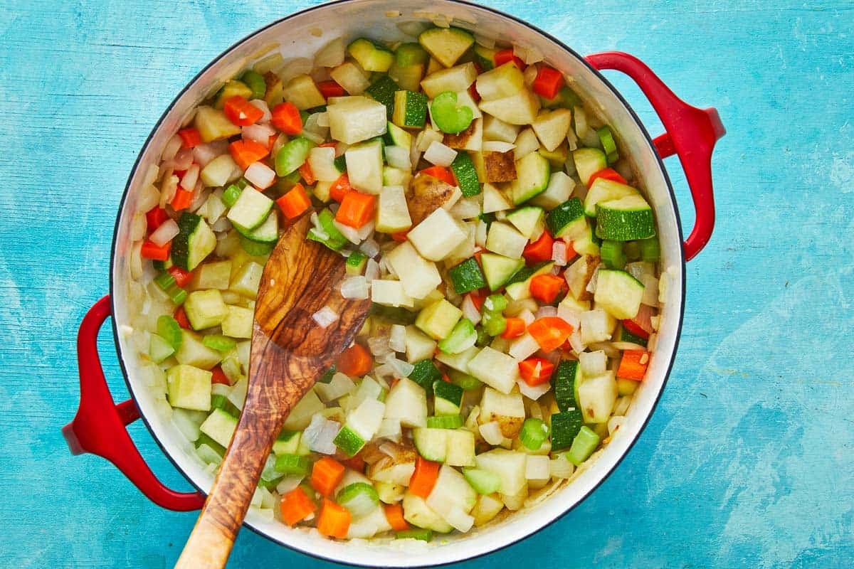 chopped carrots, celery, potatoes and zucchini cooking a a dutch oven with a wooden spoon.