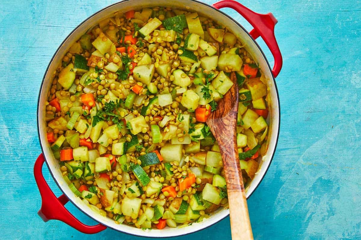 chopped carrots, celery, potatoes, zucchini and lentils cooking a a dutch oven with a wooden spoon.