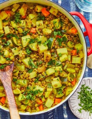 vegan lentil vegetable stew in a dutch oven with a wooden spoon, next to a bowl of chopped parsley, an empty bowl and two spoons.