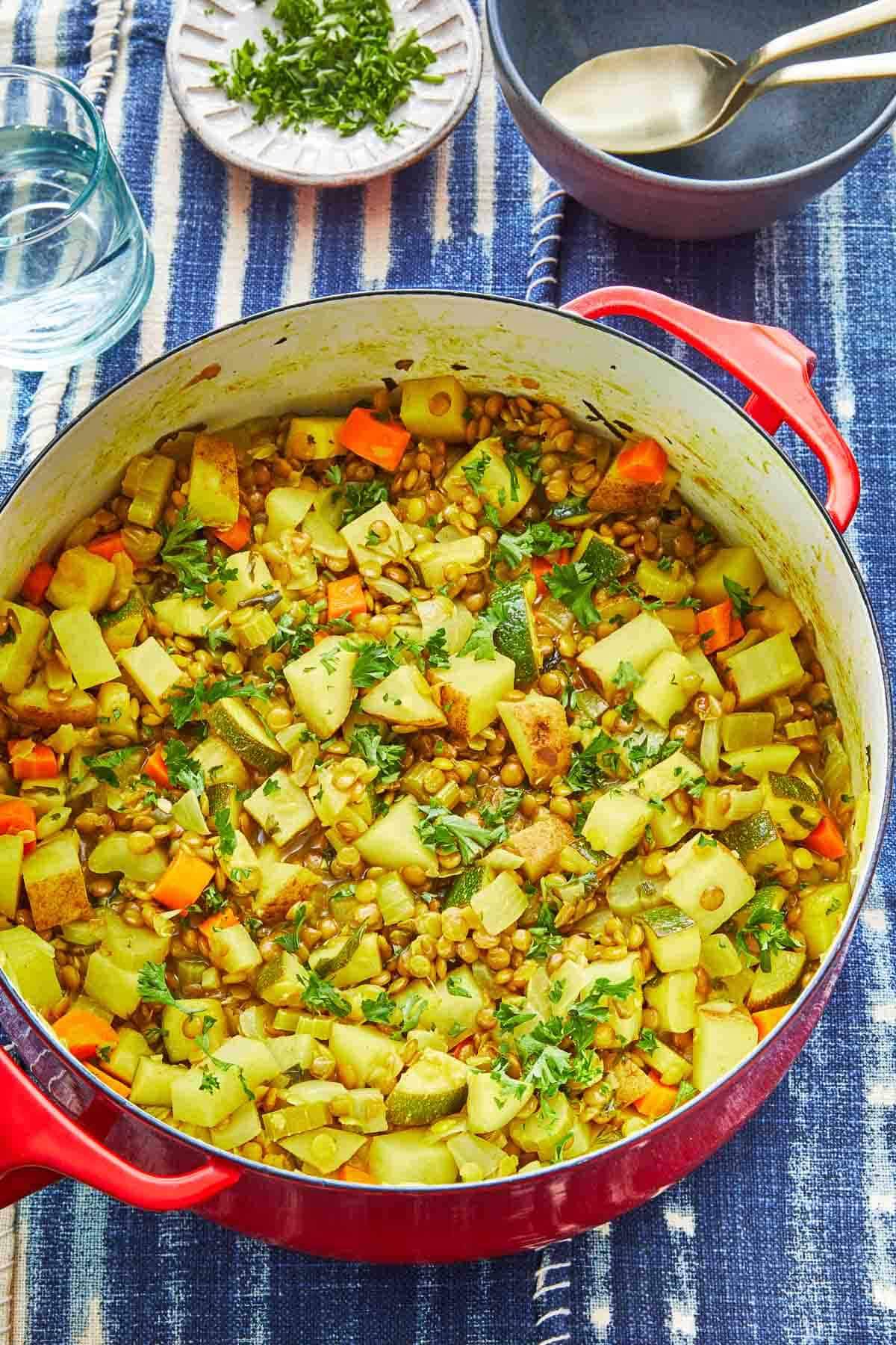 vegan lentil vegetable stew in a dutch oven with a wooden spoon, next to a glass of water, a bowl of chopped parsley and an empty bowl with two spoons.