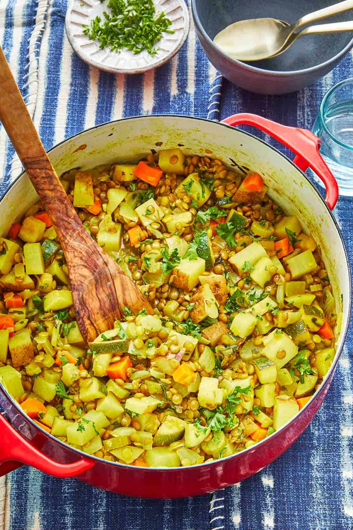 vegan lentil vegetable stew in a dutch oven with a wooden spoon, next to a bowl of chopped parsley and an empty bowl with two spoons.