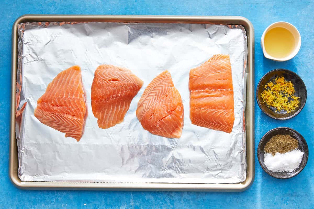 four uncooked salmon fillets on a foiled line baking sheet next to a bowl of lemon juice, a bowl of lemon zest, and a bowl of salt and pepper.