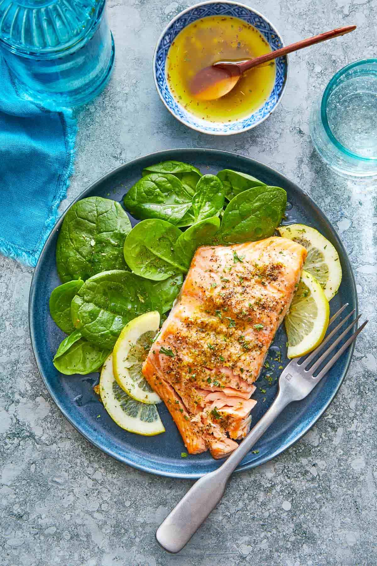 a broiled salmon fillet on a plate with lemon slices and a spinach salad with a fork next to a bowl of ladolemono dressing with a wooden spoon.