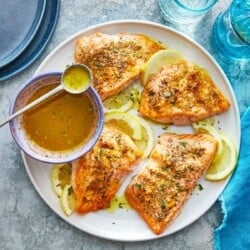 close up of four broiled salmon fillets on a plate with lemon slices and a bowl of greek ladolemono dressing with a spoon.