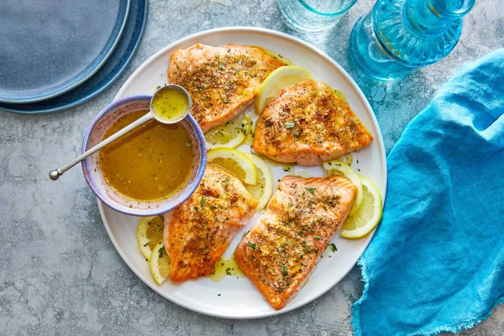 four broiled salmon fillets on a plate with lemon slices and a bowl of greek ladolemono dressing with a spoon.