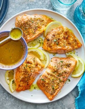 four broiled salmon fillets on a plate with lemon slices and a bowl of greek ladolemono dressing with a spoon.