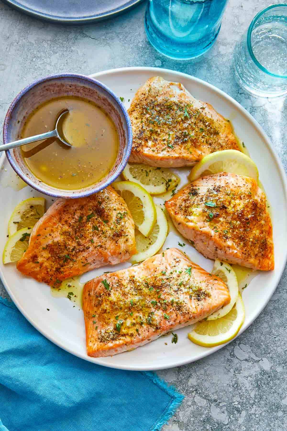 four broiled salmon fillets on a plate with lemon slices and a bowl of greek ladolemono dressing with a spoon.