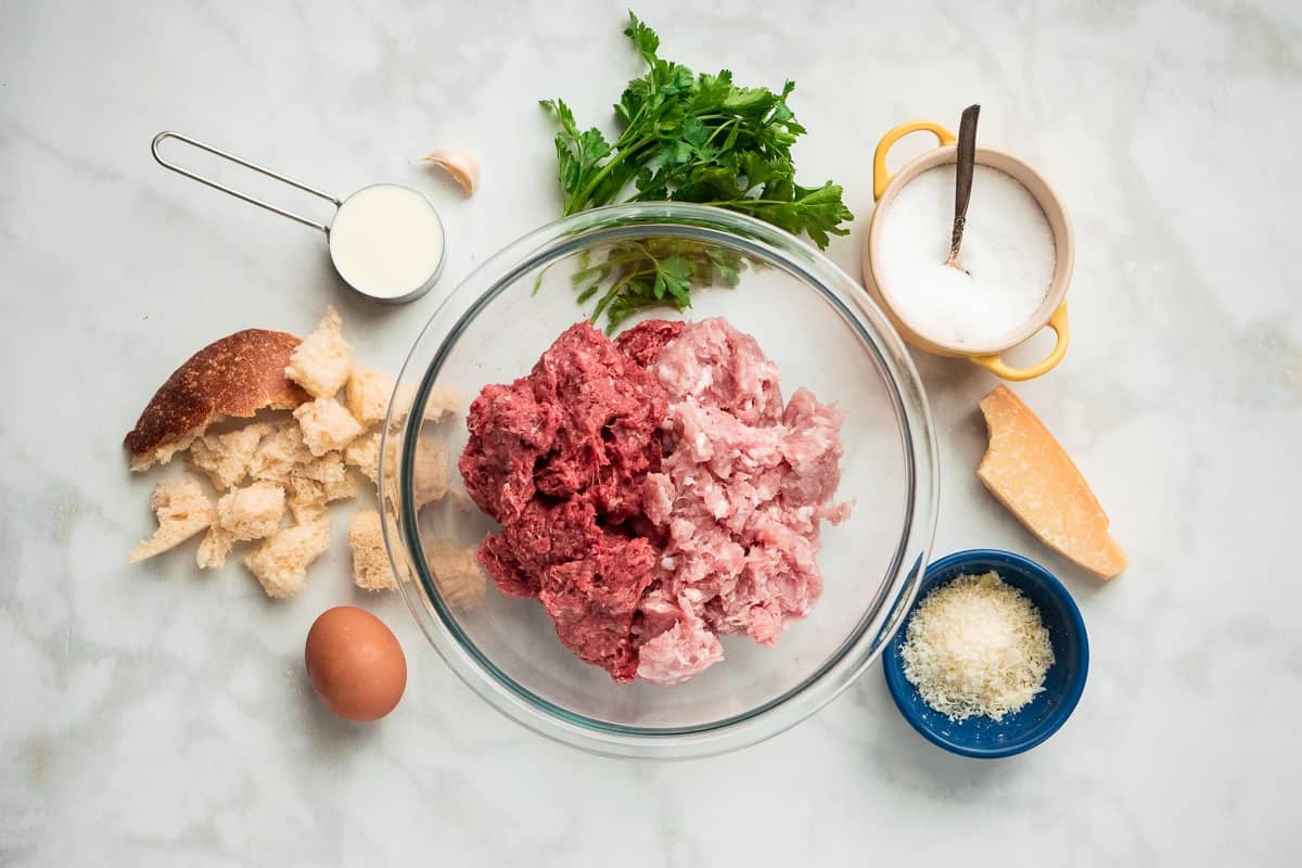 the ingredients for meatballs including bread, milk, ground beef, ground pork, an egg, parmigiano-reggiano cheese, parsley, and sea salt.