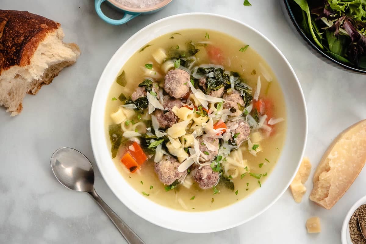 a bow of italian wedding soup next to a spoon, salad, some crusty bread and a parmesan rind.