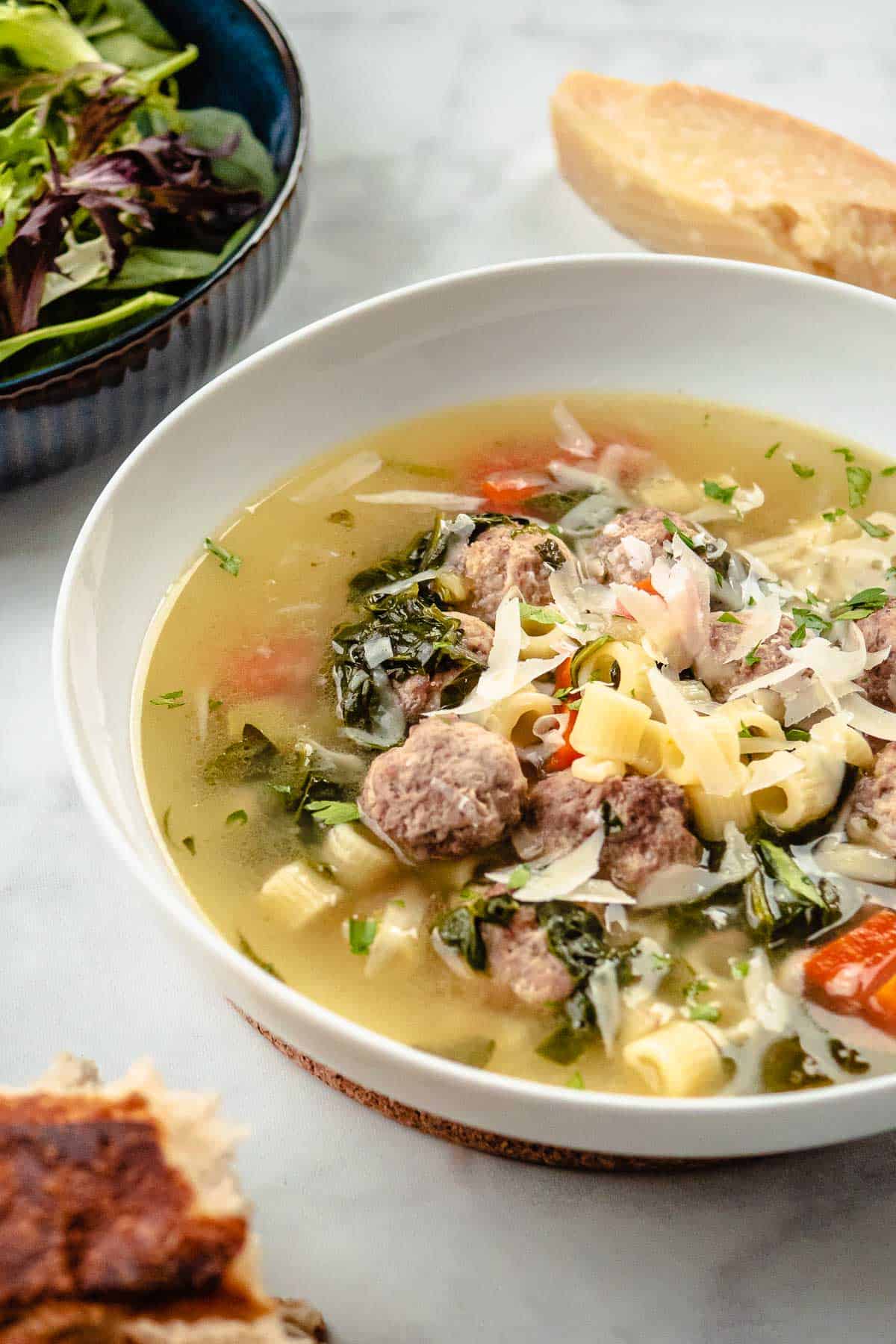 a bow of italian wedding soup next to a salad, some crusty bread and a parmesan rind.