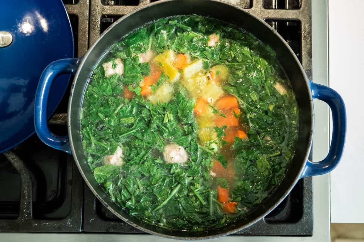 meatballs, baby spinach, carrots and celery simmering in chicken broth in a dutch oven on a stove.
