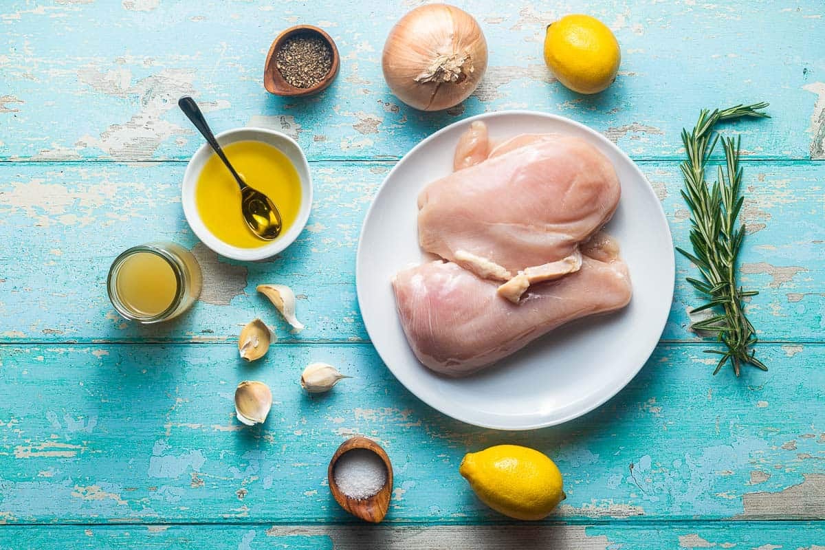 the ingredients for lemon rosemary chicken including chicken breasts on a plate, rosemary, salt, pepper, lemons, olive oil, chicken broth, garlic, and an onion.