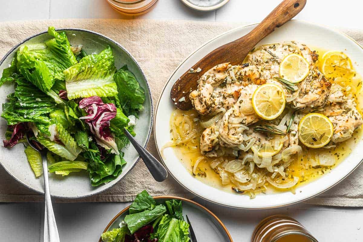 lemon rosemary chicken topped with lemon slices on a plate with a wooden spoon next to a salad in a bowl.