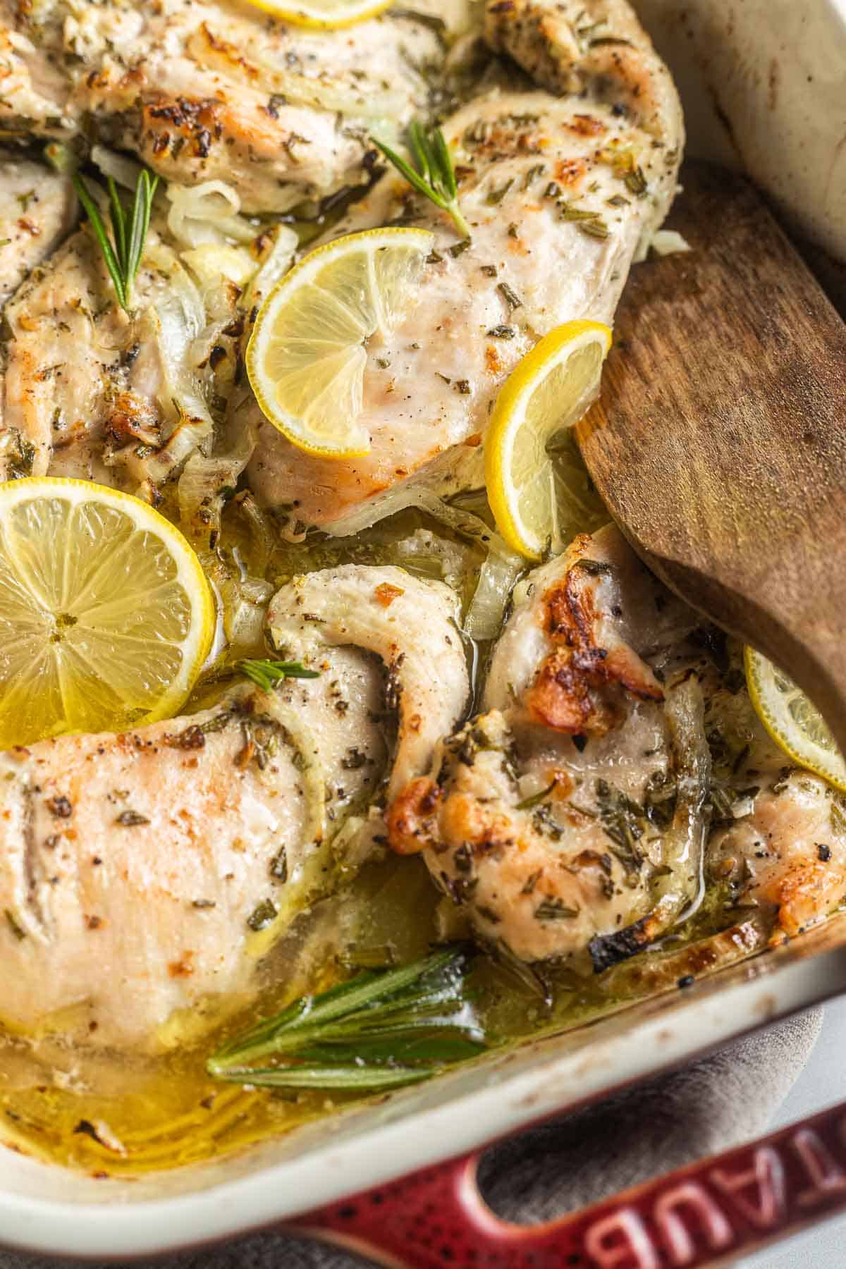 a close up of cooked rosemary chicken in a baking dish with lemon slices and a wooden spoon.