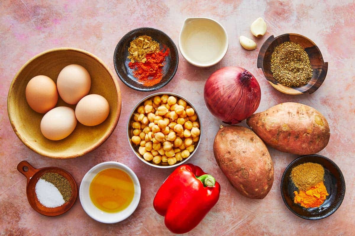 ingredients for sweet potato hash including eggs, onion, sweet potatoes, chickpeas, garlic, red bell pepper, extra virgin olive oil, white vinegar, and spices.