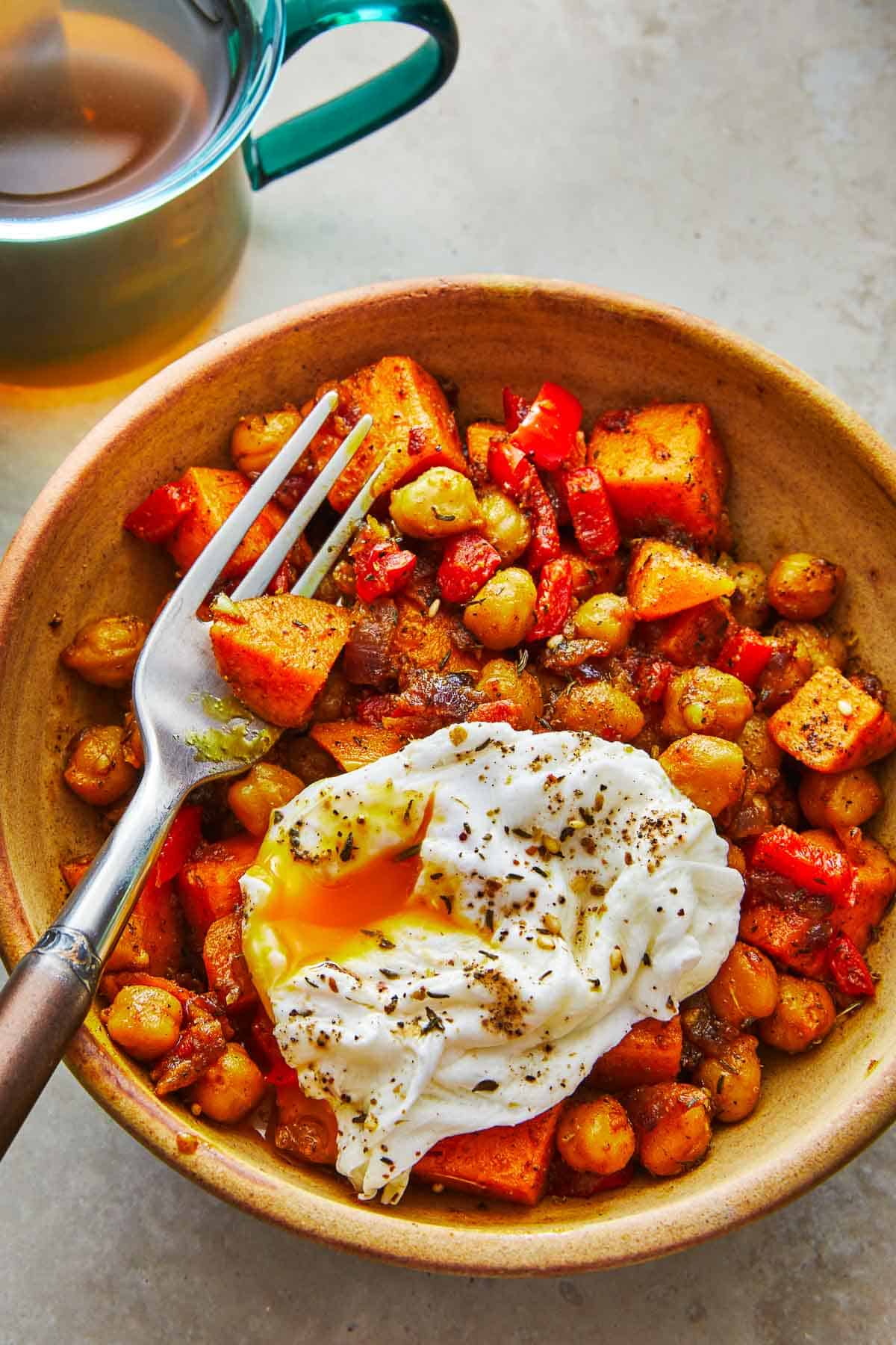 a bowl of sweet potato hash with a poached egg and a fork next to a mug of tea.