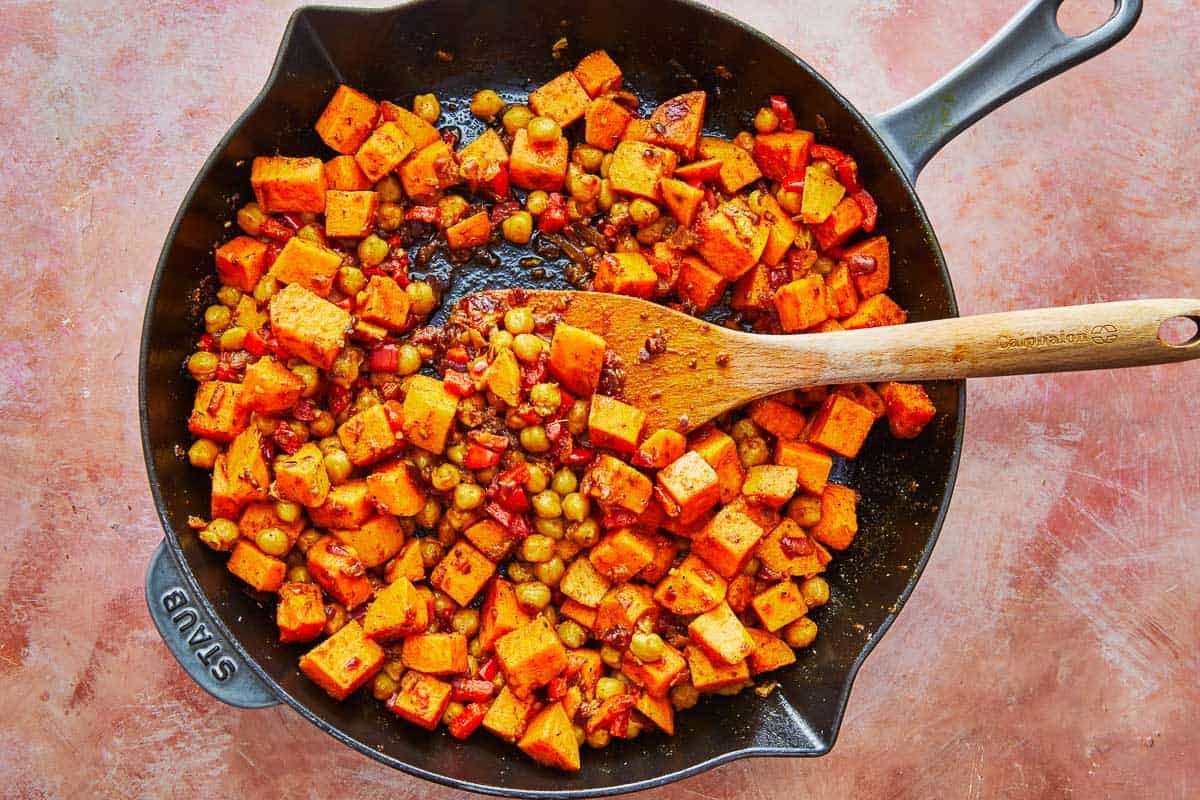 red onion, sweet potatoes and chickpeas being sauteed in a cast iron skillet with a wooden spoon.