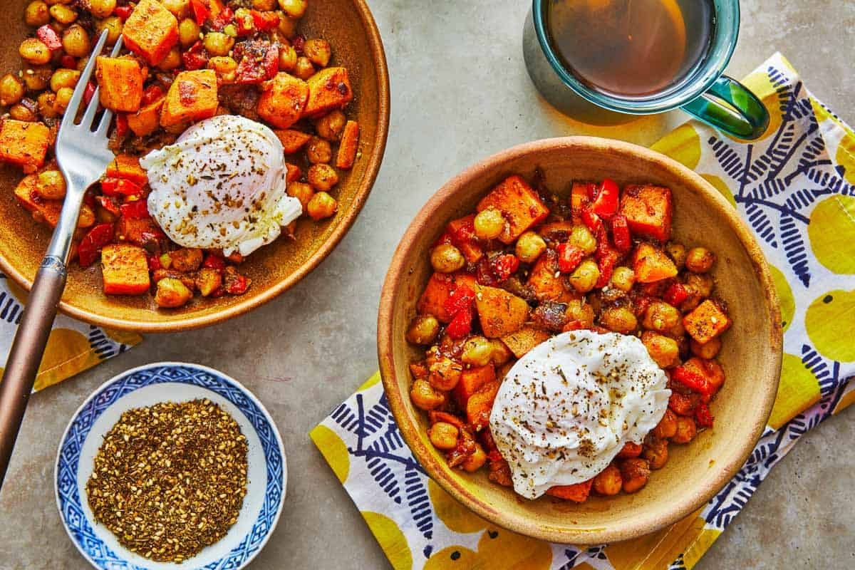 two bowls of sweet potato hash with poached eggs next to a bowl of za'atar and a mug of tea.