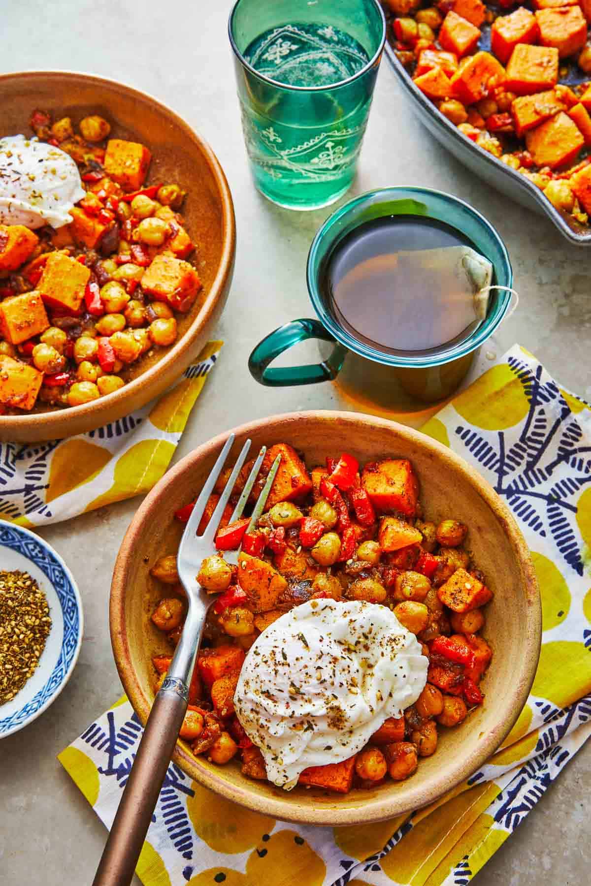 a bowl of sweet potato has with a poached egg and a fork next to a cup of tea and a glass of water.