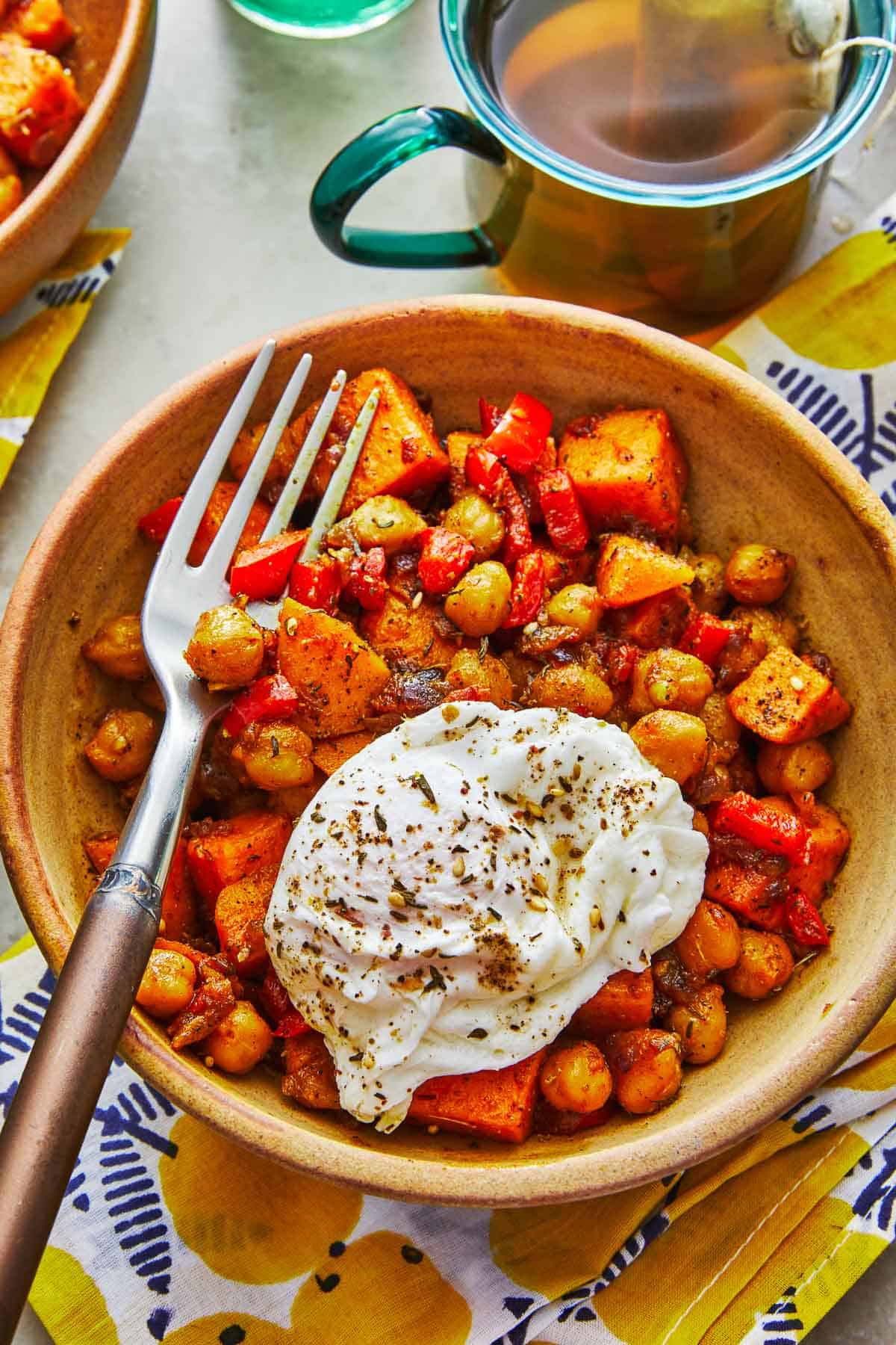 a bowl of sweet potato hash with a poached egg and a fork next to a mug of tea.