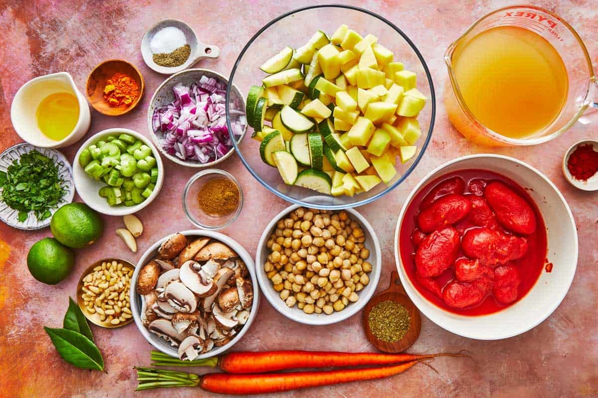 ingredients for homemade vegetable soup including olive oil, mushrooms, onion, parsley, celery, zucchini, potatoes, tomatoes, chickpeas, carrots, bay leaves, broth, lime, pine nuts, garlic and spices.