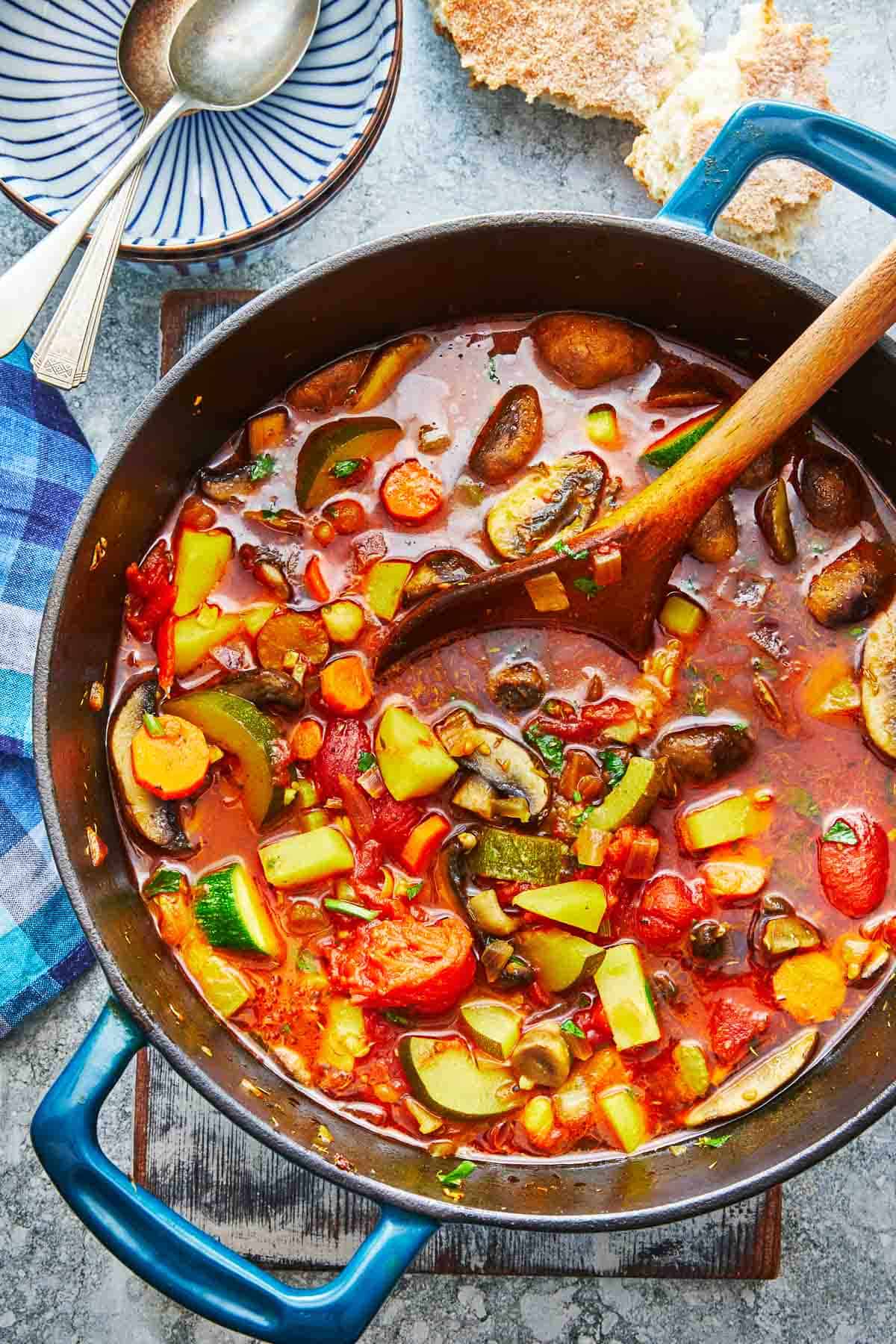 homemade vegetable soup in a dutch oven with a wooden spoon next to a bowl with two silver spoons.