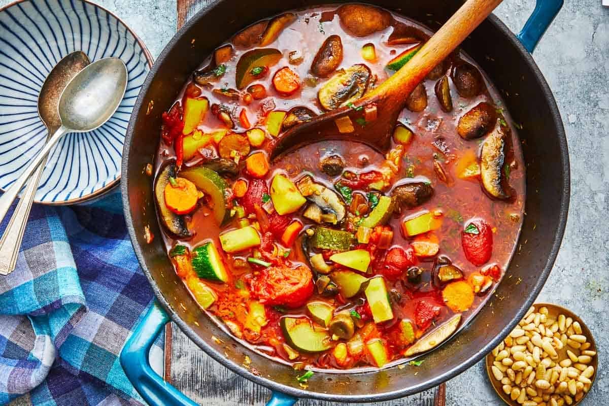 homemade vegetable soup in a dutch oven with a wooden spoon next to a bowl with two silver spoons.