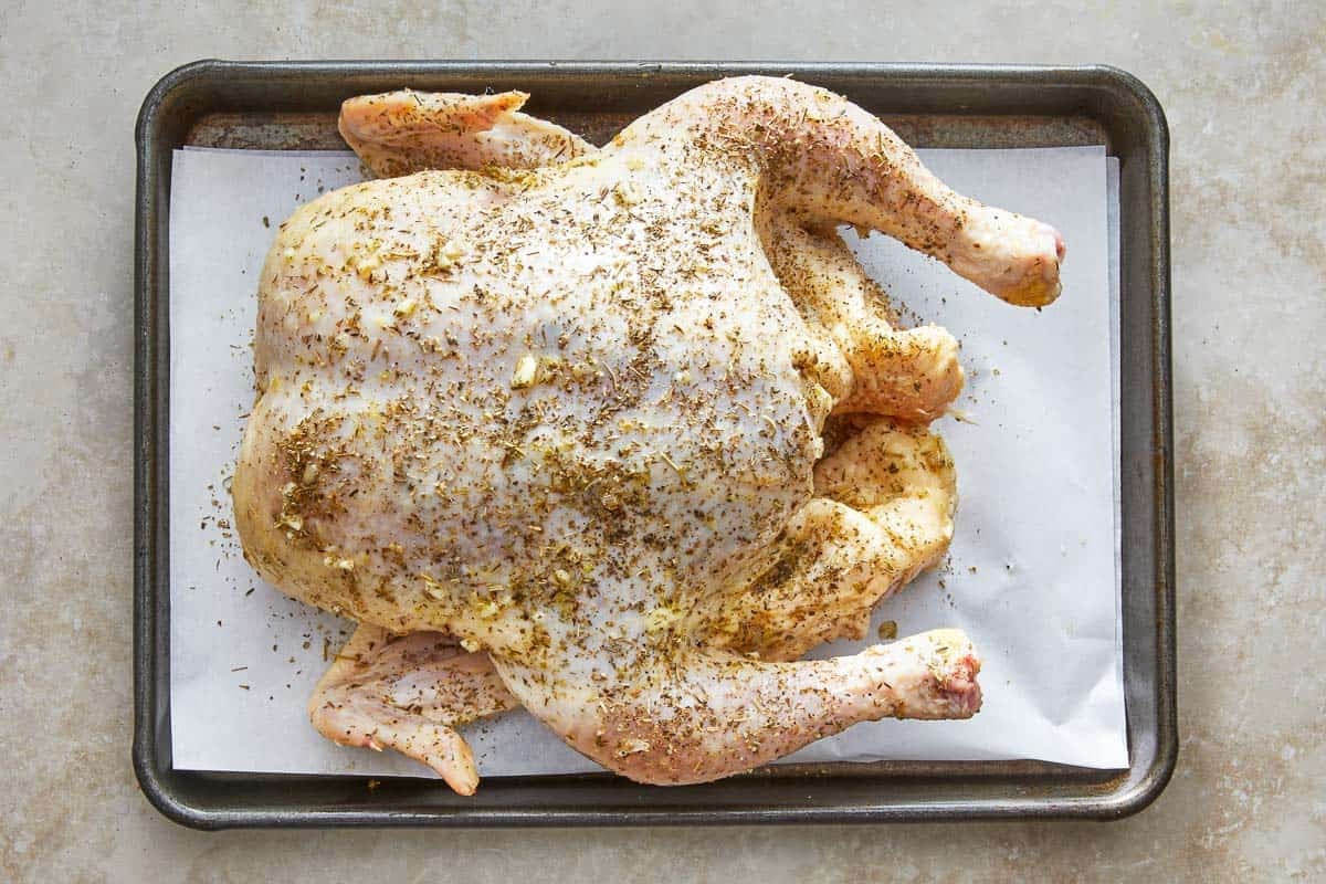 a whole uncooked chicken with an italian seasoning rub on a parchment lined baking sheet.