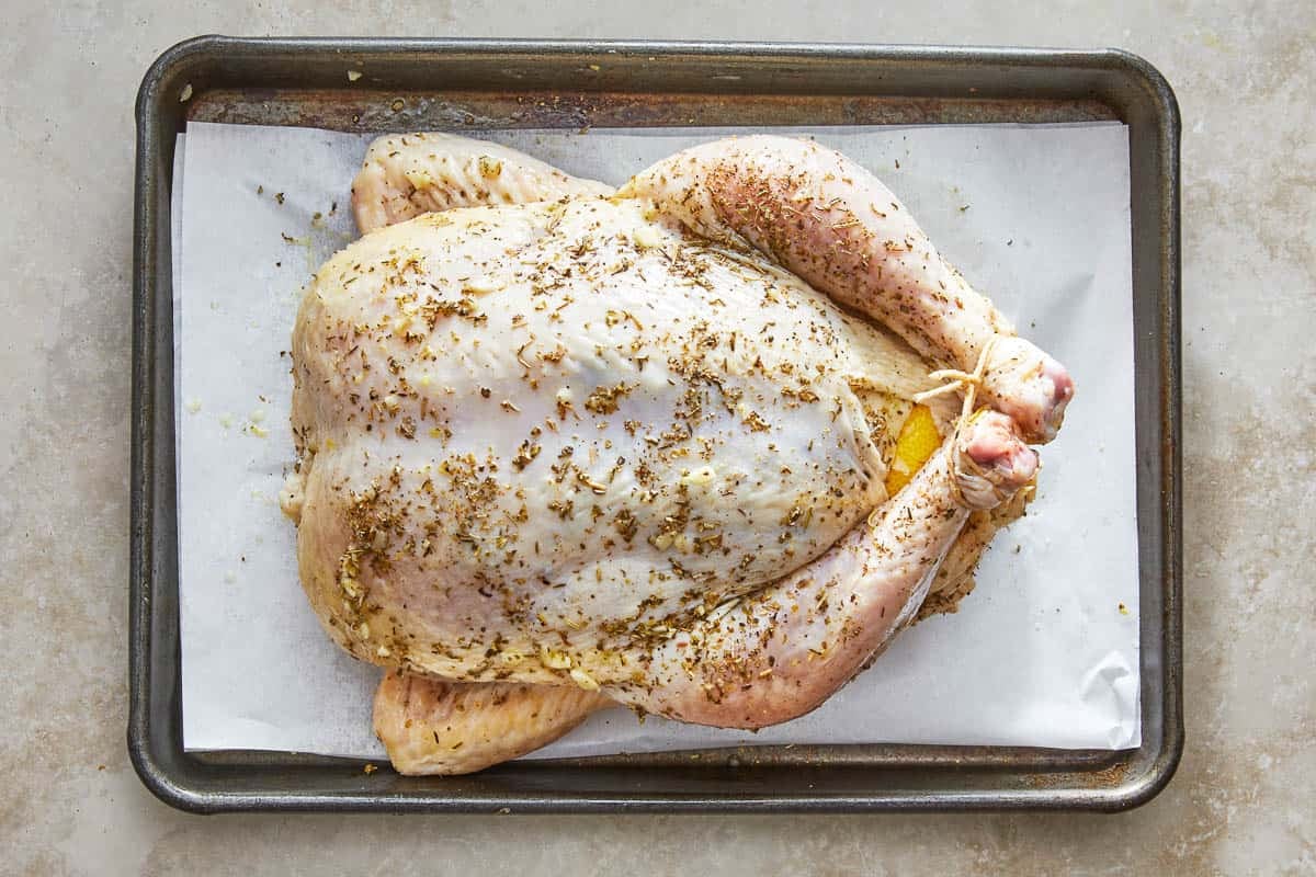 a whole uncooked chicken with an italian seasoning rub trussed on a parchment lined baking sheet.