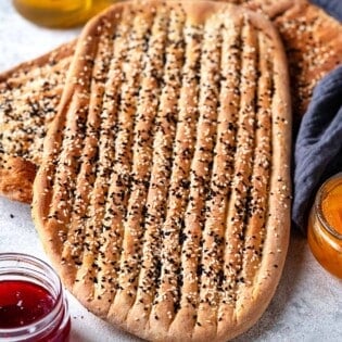 two pieces of barbari bread surrounded by bowls of toppings such as feta cheese, honey, and jam.