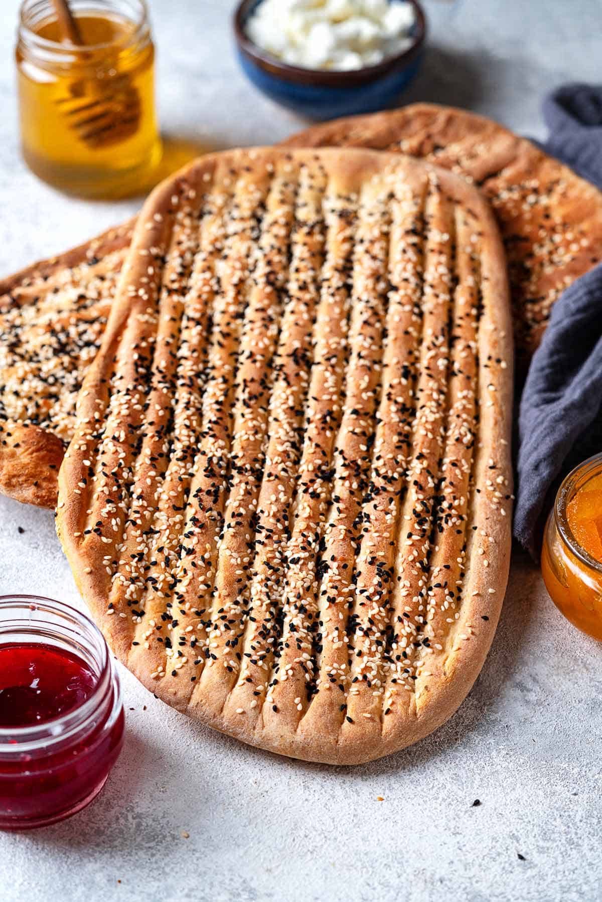 two pieces of barbari bread surrounded by bowls of toppings such as feta cheese, honey, and jam.