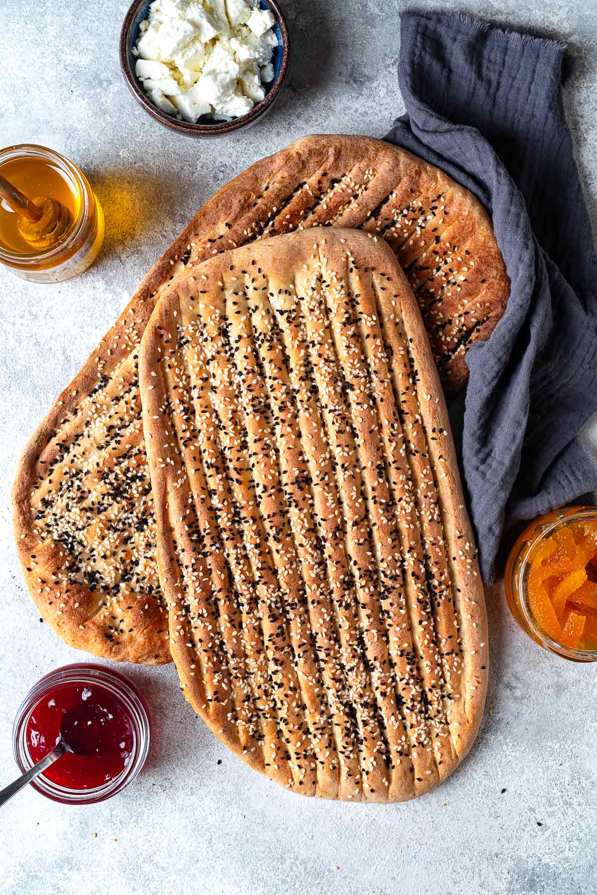 two pieces of barbari bread surrounded by bowls of toppings such as feta cheese, honey, and jam.