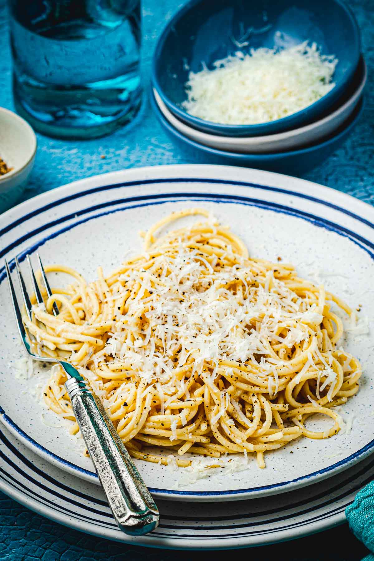 Cacio e pepe on a plate with a fork next to a bowl of grated Pecorino Romano cheese.