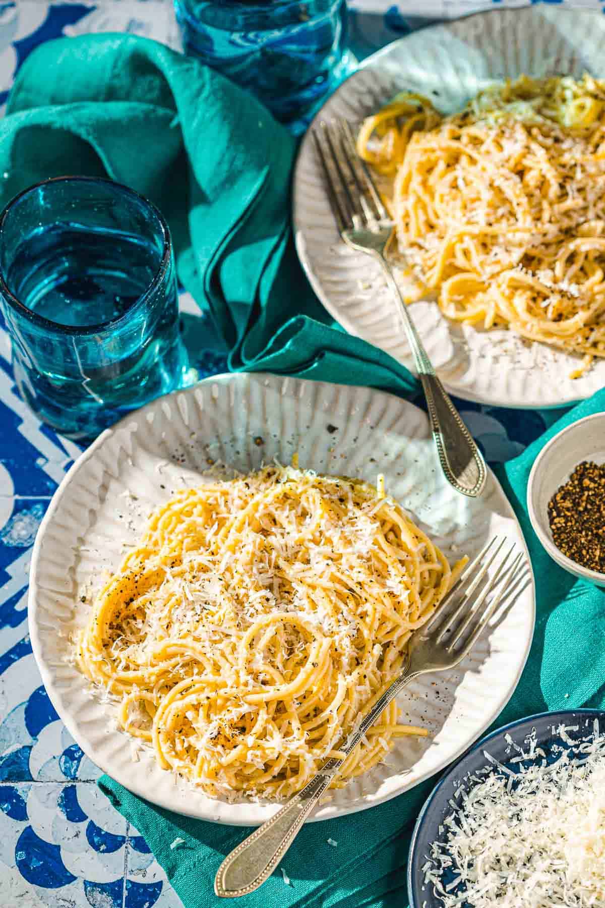 two plates of cacio e pepe, each with a fork, next to a glass of water and a bowl of pepper.