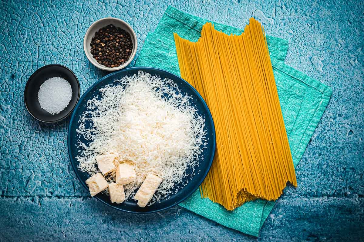 ingredients for cacio e pepe including salt, peppercorns, grated Pecorino Romano cheese, and spaghetti.