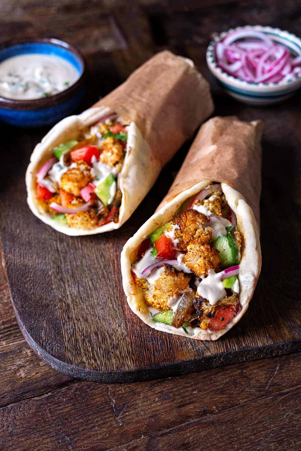 two cauliflower shawarma pitas with sides of tahini sauce and sliced red onion in bowls.