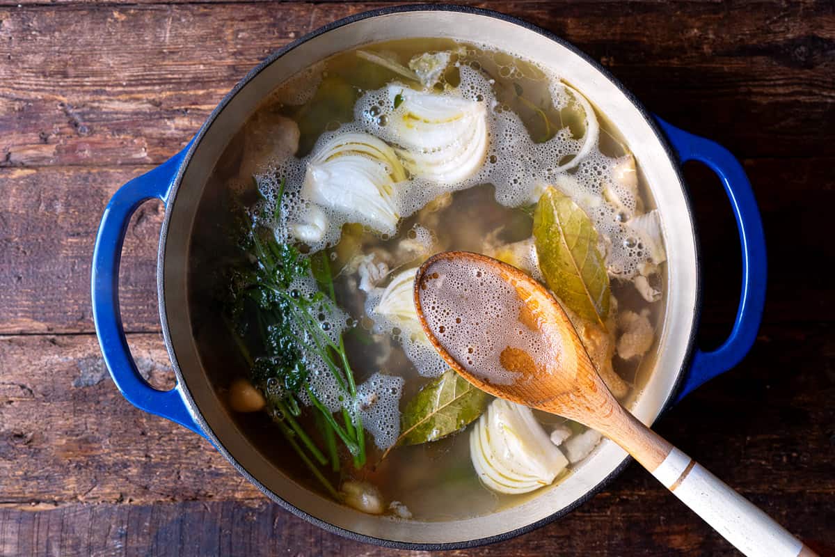 chopped celery, carrots, onion, garlic, thyme, bay leaves parsley stems and a chicken carcass covered with water in a stock pot with a wooden spoon.