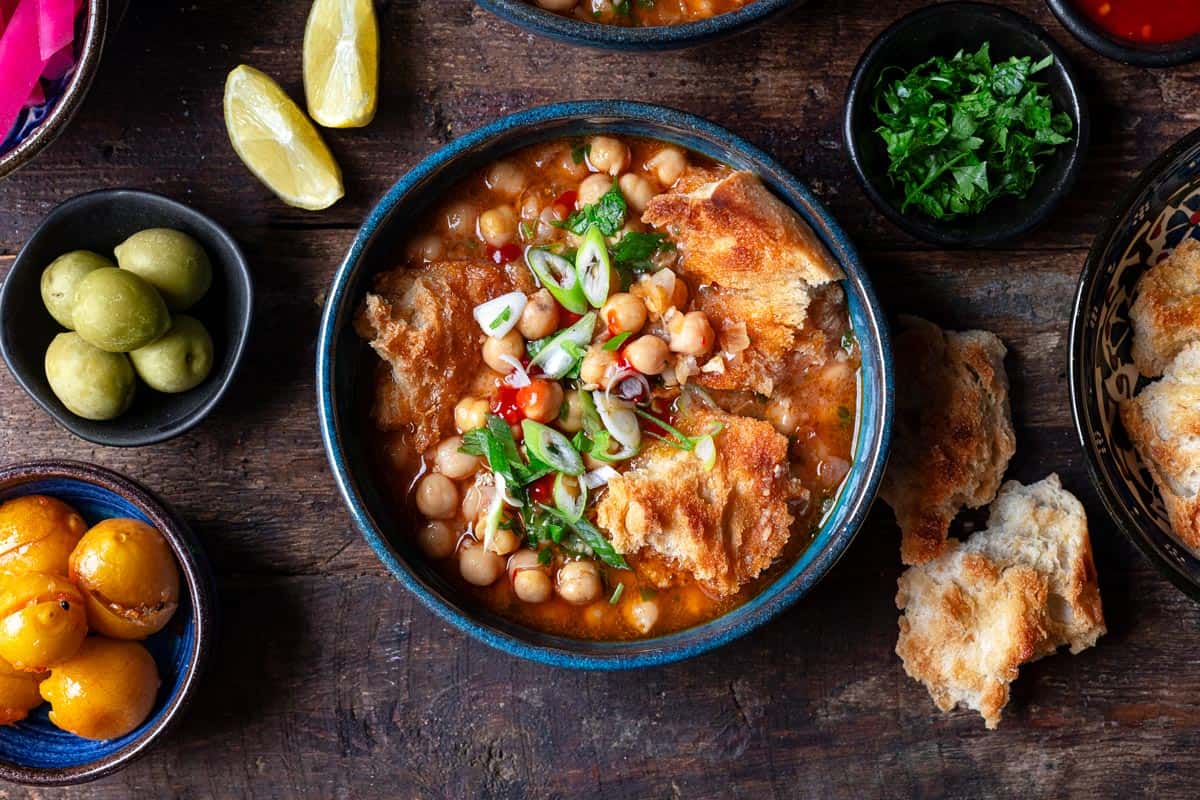 lablabi tunisian chickpea stew in a bowl, next to bowls of various toppings and torn up toasted bread.