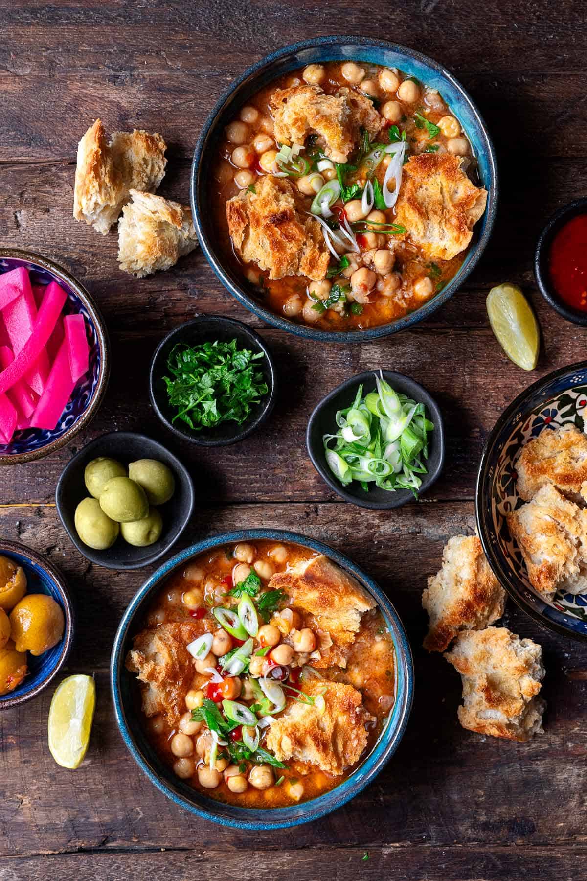 two bowls of lablabi tunisian chickpea stew next to bowls of various toppings and torn up toasted bread..