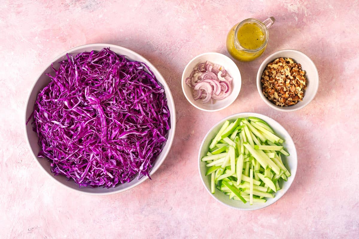 ingredients for red cabbage salad including apple cider vinaigrette, shredded red cabbage, julienned apples, sliced shallots, and chopped walnuts.