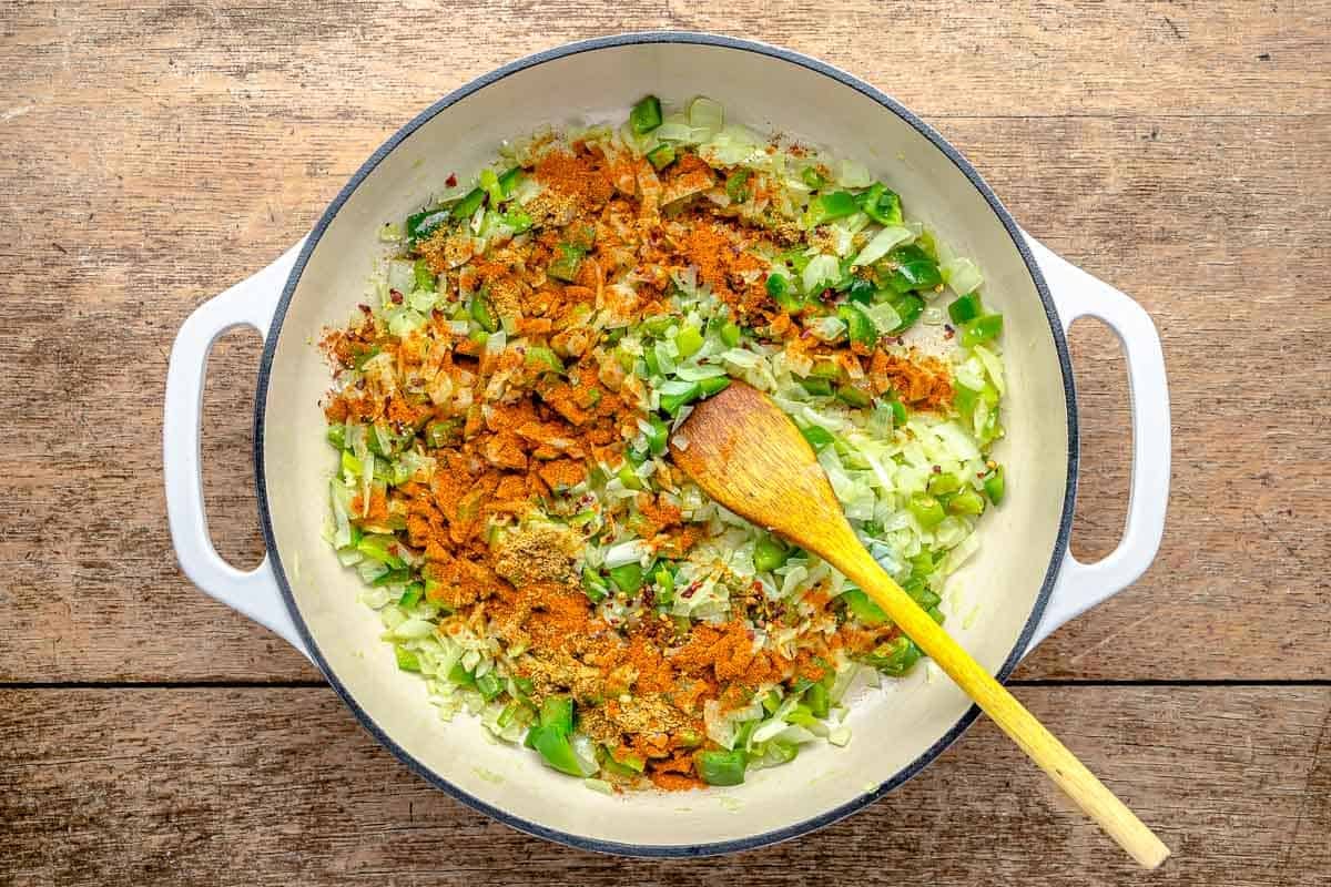 chopped yellow onions and green peppers with a sprinkle of spices being sauteed in a pan with a wooden spoon.