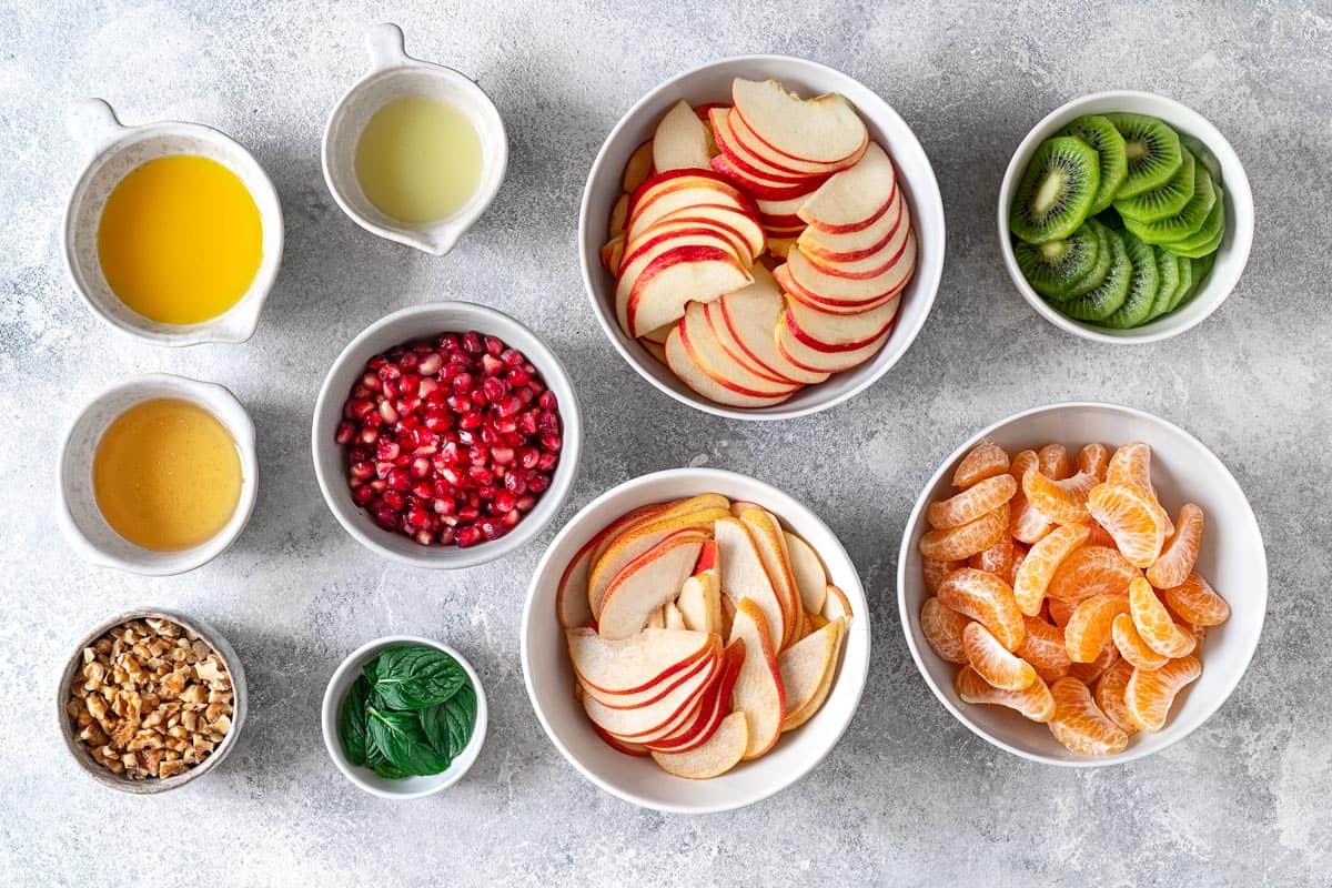 ingredients for winter fruit salad included sliced apples, oranges, kiwi, pear, pomegranate arils, mint, walnuts, honey, orange juice and lime juice.
