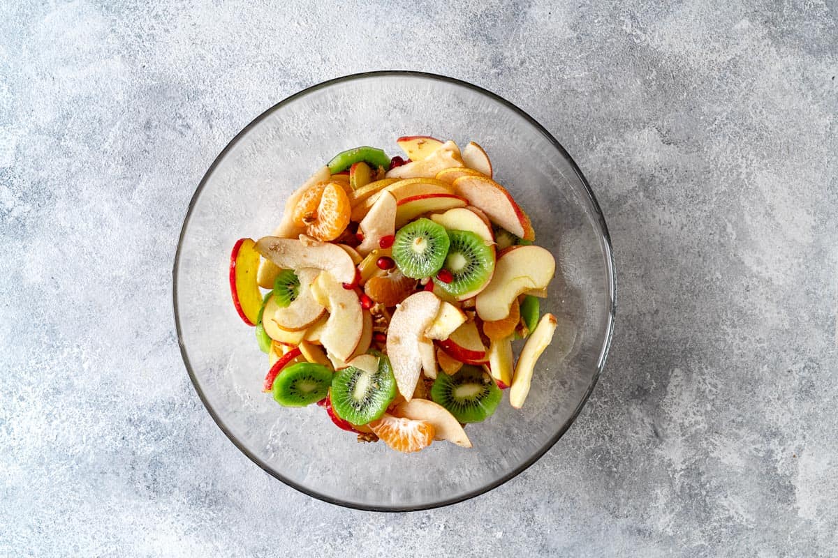 sliced kiwi, apples, pears, oranges and pomegranate arils in a mixing bowl.