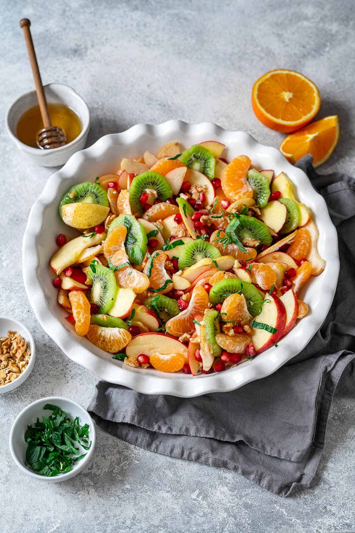 winter fruit salad in a bowl next to small bowls of chopped walnut, honey, chopped mint and orange wedges.