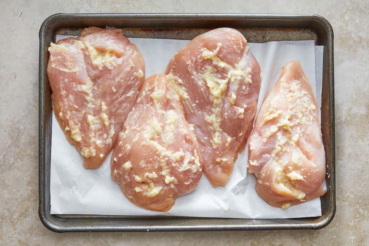 4 raw chicken breasts rubbed with garlic on a parchment-lined baking sheet.